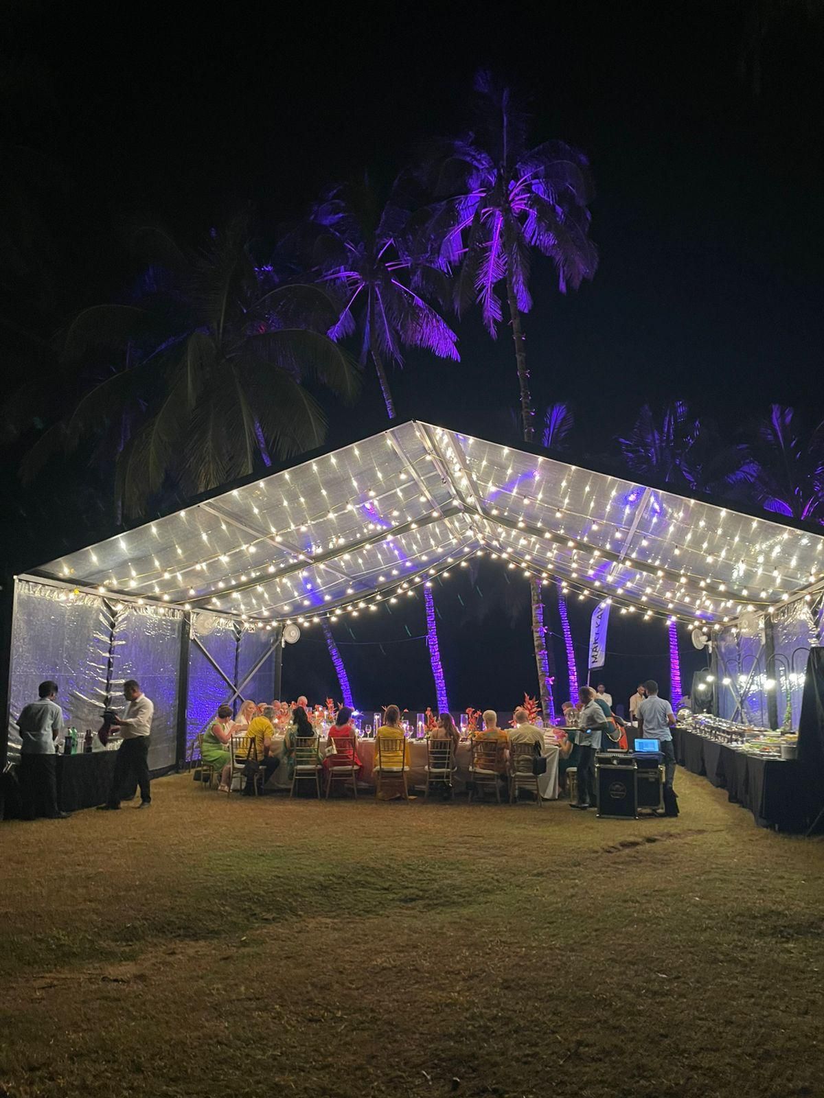 A clear marquee lit with string lights for a group dinner under palm trees