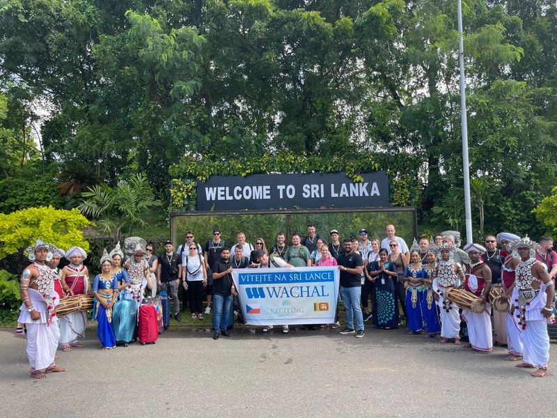 A large Czech tour group holding a welcome banner under the Welcome to Sri Lanka sign, flanked by drummers and dancers