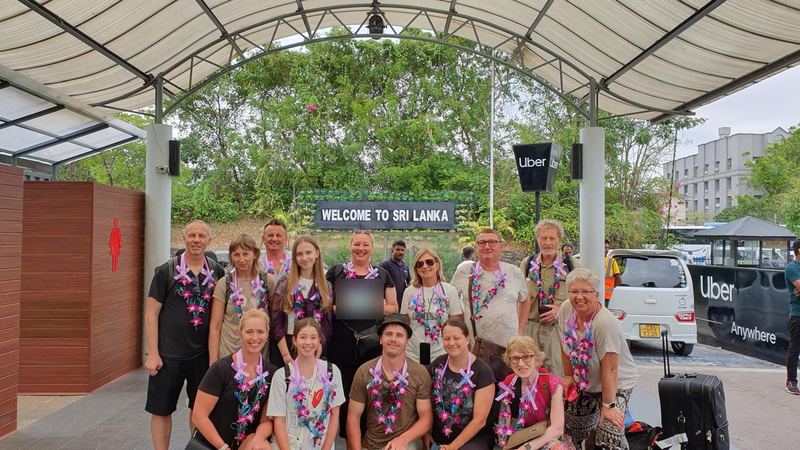 A group with orchid garlands outside the airport under the welcome sign